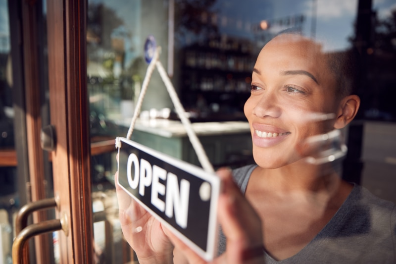 Female Owner Of Start Up Coffee Shop Or Restaurant Turning Round Open Sign On Door.