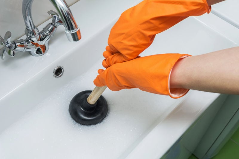 Summertime plumbing problems to avoid. Person with orange gloves on trying to unclog a drain with a plunger.