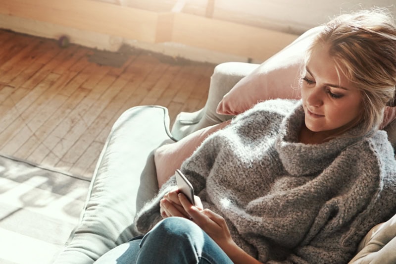 Expert Tips: How to Keep Your Older Furnace Running for Years! Photo of a woman in a sweater playing on her phone on the couch.