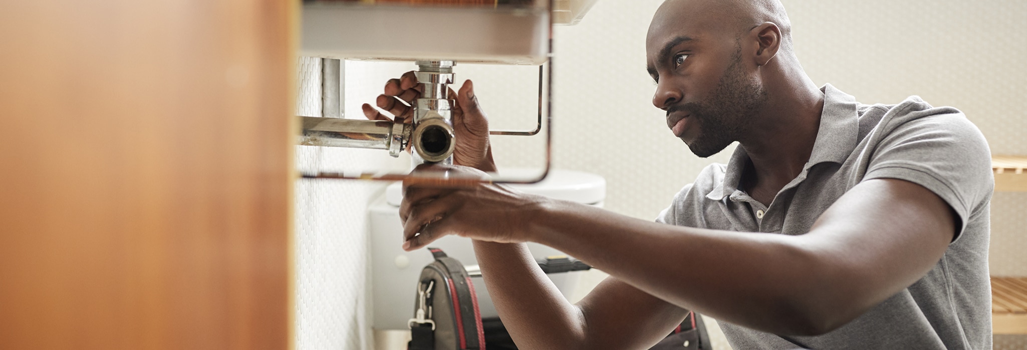 Young black male plumber sitting on the floor fixing a bathroom sink, seen from doorway