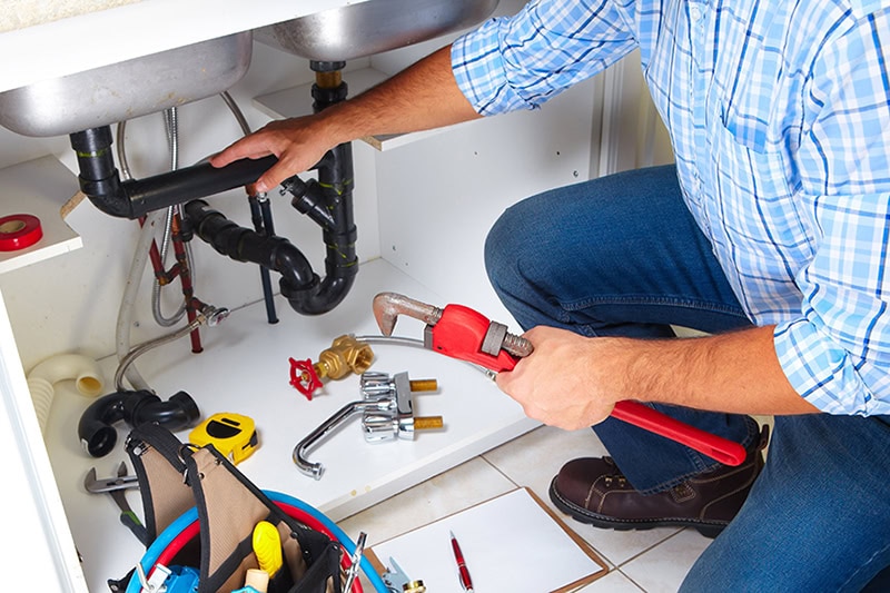 Plumbing Tips for Hosting Guests Over the Holidays. Photo of a plumber performing maintenance under a sink.
